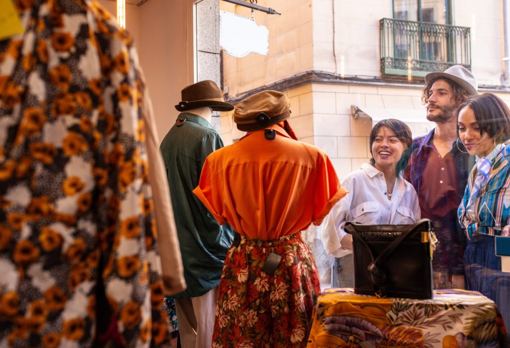 Friends looking at clothes behind shop window Young man and women smiling and looking at clothes inside small store while standing on street near display window